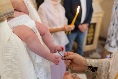Close-up of priest's hands anointing baby's legs during Orthodox baptismal ceremony. Focus on child,...