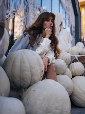Fashionable woman in white knit dress and gray jacket sitting among large white pumpkins and ghostly figures, looking away. Halloween mood.の写真素材