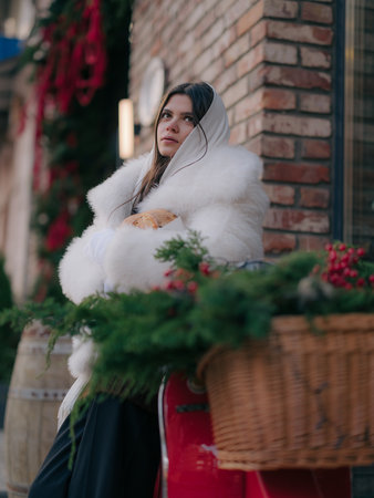 Young woman in white winter coat and mittens sitting on vintage red scooter with festive Christmas decorated wicker basket, holding baked roll on street.の写真素材
