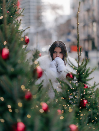 Close up portrait of thoughtful young woman in white winter coat and mittens, looking intently at camera, framed by festive Christmas tree branches.の写真素材