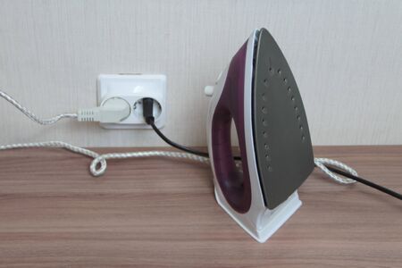A lilac electric iron stands on a table plugged into a wall outlet.