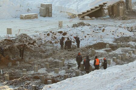 Construction workers in overalls in orange helmets work at a construction site among piles in winter...