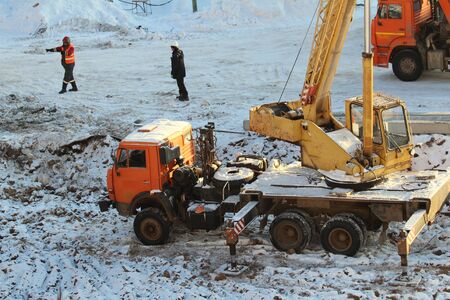 Truck crane truck with orange and yellow cabs with extended supports works at a construction site against the background of construction workers and snow in winter in Russia.の写真素材