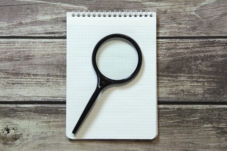 Notepad with a blank white sheet in a checker paper lies with magnifying glass on the background of wooden boards.の写真素材