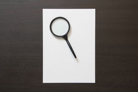 Template of white paper with magnefy on dark wenge color wooden background. Concept of searching, detecting and solving problems. Stock photo with empty space for text and design.の写真素材