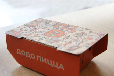 08-02-2020, Russia, Syktyvkar. Orange cardboard company box for Dodo snacks with a side inscription pizza and a corporate design and logo on the lid is standing on a table in a restaurant near the window.のeditorial素材