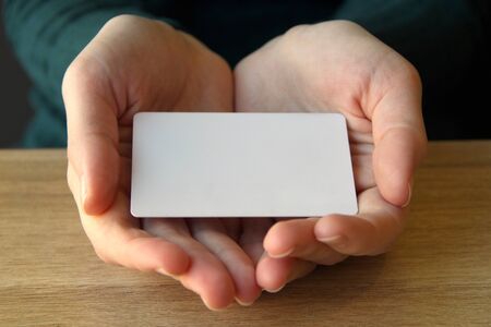 A white rectangular card lies in the opened palms of a girl in a blue sweater. Mockup for design,...