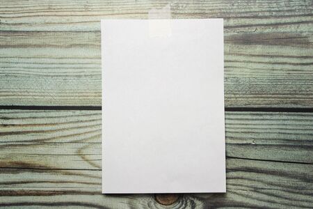 A white sheet of A4 paper hanging on a wall glued with a piece of paper tape on a wooden background.の写真素材