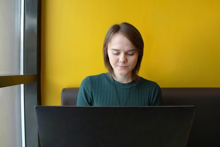Beautiful young girl sitting in a cafe at a table with a laptop. Works at the computer and types on the keyboard. Looks at the screen with an emotional expression.の写真素材
