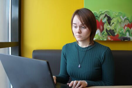 Beautiful young girl sitting in a cafe at a table with a laptop. Works at the computer and types on the keyboard. Looks at the screen with an emotional expression.の写真素材