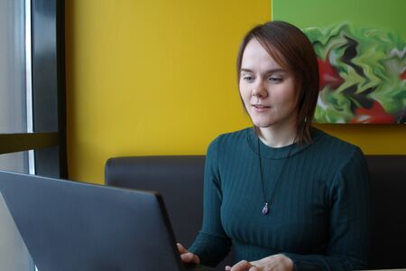 Beautiful young girl sitting in a cafe at a table with a laptop. Works at the computer and types on the keyboard. Looks at the screen with an emotional expression.の写真素材