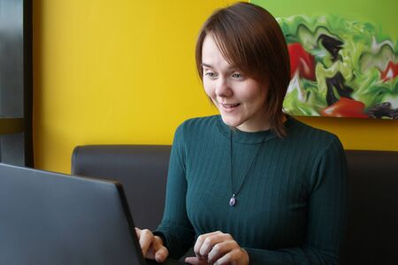 Girl with a perplexed surprised tense expression sits sitting working at a laptop at a table in a cafe on a brown sofa against a yellow wall.の写真素材