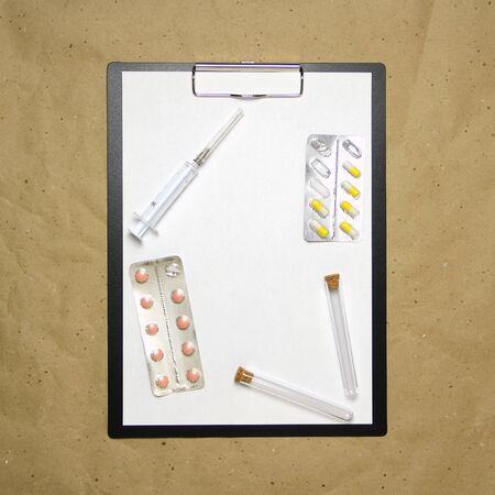 The tablet with a clip for the clip of paper with a white sheet a4 lies against the background of craft brown crumpled paper. On top is a medical syringe, pills in a blister and two transparent empty tubes with corks. Place for text and layout for design.の写真素材