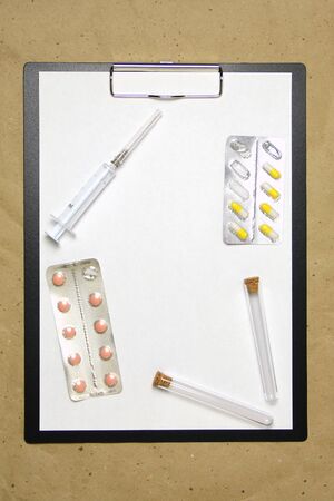 The tablet with a clip for the clip of paper with a white sheet a4 lies against the background of craft brown crumpled paper. On top is a medical syringe, pills in a blister and two transparent empty tubes with corks. Place for text and layout for design.の写真素材