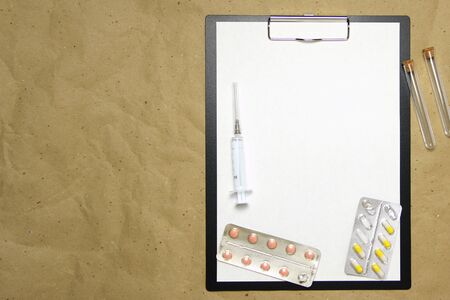 The tablet with a clip for the clip of paper with a white sheet a4 lies against the background of craft brown crumpled paper. On top is a medical syringe, pills in a blister and two transparent empty tubes with corks. Place for text and layout for design.の写真素材