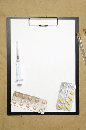 The tablet with a clip for the clip of paper with a white sheet a4 lies against the background of craft brown crumpled paper. On top is a medical syringe, pills in a blister and two transparent empty tubes with corks. Place for text and layout for design.の写真素材