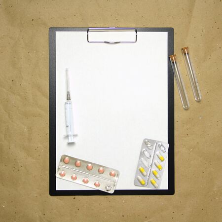 The tablet with a clip for the clip of paper with a white sheet a4 lies against the background of craft brown crumpled paper. On top is a medical syringe, pills in a blister and two transparent empty tubes with corks. Place for text and layout for design.の写真素材
