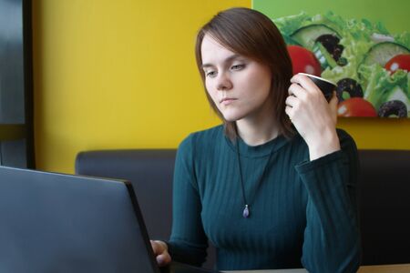 A girl with a pensive calm expression on her face sits at a laptop at a table in a cafe on a brown sofa against a yellow wall. He is looking at a computer monitor, holding a paper cup with coffee in his hand.の写真素材