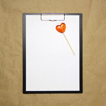 The tablet with a clip for the clip of paper with a white sheet a4 lies against the background of craft brown crumpled paper. On top lies a caramel lollipop of orange red in the shape of a heart. Place for text and layout for design.の写真素材