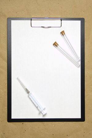 The tablet with a clip for the clip of paper with a white sheet a4 lies against the background of craft brown crumpled paper. On top is a medical syringe and two transparent empty tubes with stoppers. Place for text and layout for design.の写真素材