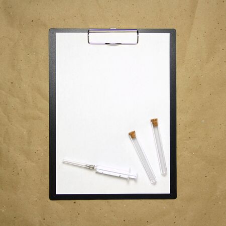 The tablet with a clip for the clip of paper with a white sheet a4 lies against the background of craft brown crumpled paper. On top is a medical syringe and two transparent empty tubes with stoppers. Place for text and layout for design.の写真素材