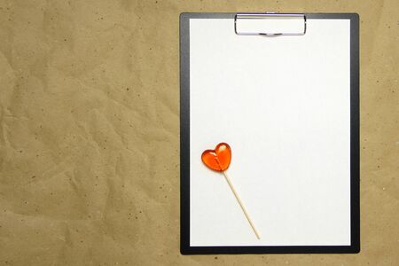 The tablet with a clip for the clip of paper with a white sheet a4 lies against the background of craft brown crumpled paper. On top lies a caramel lollipop of orange red in the shape of a heart. Place for text and layout for design.の写真素材