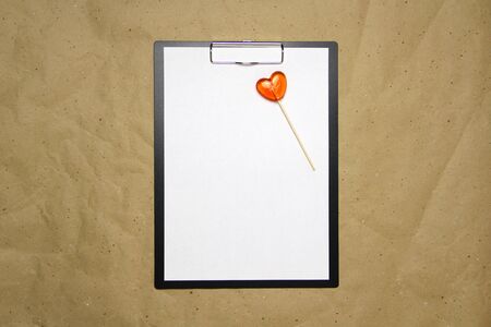 The tablet with a clip for the clip of paper with a white sheet a4 lies against the background of craft brown crumpled paper. On top lies a caramel lollipop of orange red in the shape of a heart. Place for text and layout for design.の写真素材