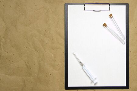 The tablet with a clip for the clip of paper with a white sheet a4 lies against the background of craft brown crumpled paper. On top is a medical syringe and two transparent empty tubes with stoppers. Place for text and layout for design.の写真素材