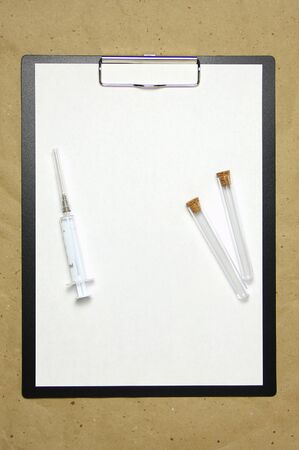 The tablet with a clip for the clip of paper with a white sheet a4 lies against the background of craft brown crumpled paper. On top is a medical syringe and two transparent empty tubes with stoppers. Place for text and layout for design.の写真素材