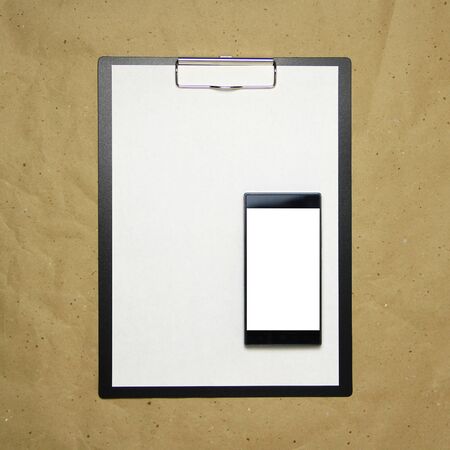 The tablet with a clip for the clip of paper with a white sheet a4 lies against the background of craft brown crumpled paper. Above is a mobile phone with a white screen. Place for text and layout for design.の写真素材