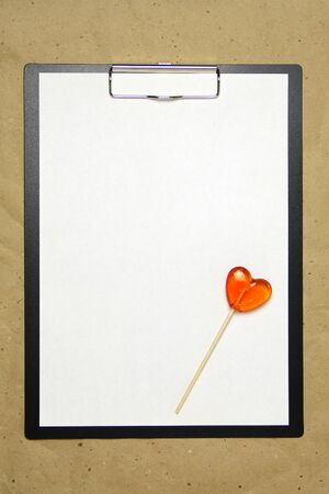 The tablet with a clip for the clip of paper with a white sheet a4 lies against the background of craft brown crumpled paper. On top lies a caramel lollipop of orange red in the shape of a heart. Place for text and layout for design.の写真素材