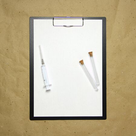 The tablet with a clip for the clip of paper with a white sheet a4 lies against the background of craft brown crumpled paper. On top is a medical syringe and two transparent empty tubes with stoppers. Place for text and layout for design.の写真素材