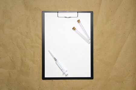 The tablet with a clip for the clip of paper with a white sheet a4 lies against the background of craft brown crumpled paper. On top is a medical syringe and two transparent empty tubes with stoppers. Place for text and layout for design.の写真素材