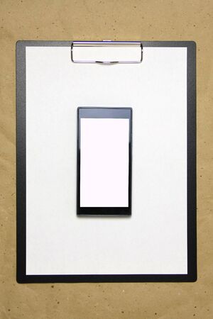 The tablet with a clip for the clip of paper with a white sheet a4 lies against the background of craft brown crumpled paper. Above is a mobile phone with a white screen. Place for text and layout for design.の写真素材