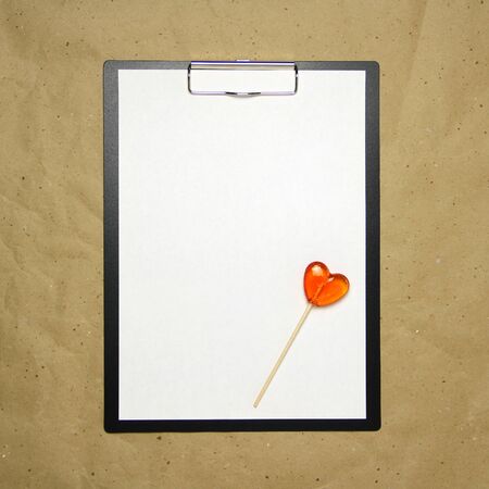 The tablet with a clip for the clip of paper with a white sheet a4 lies against the background of craft brown crumpled paper. On top lies a caramel lollipop of orange red in the shape of a heart. Place for text and layout for design.の写真素材