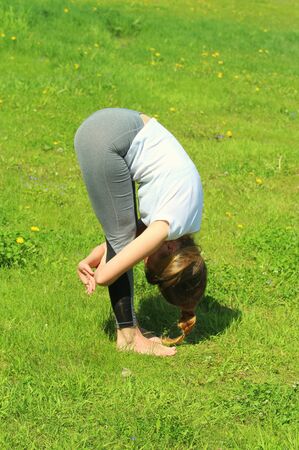 Young woman of European appearance does yoga in summer nature. Woman standing in forward tilt pose with hand lock, baddha hasta uttanasana. High quality photo for web and print with empty space designの写真素材