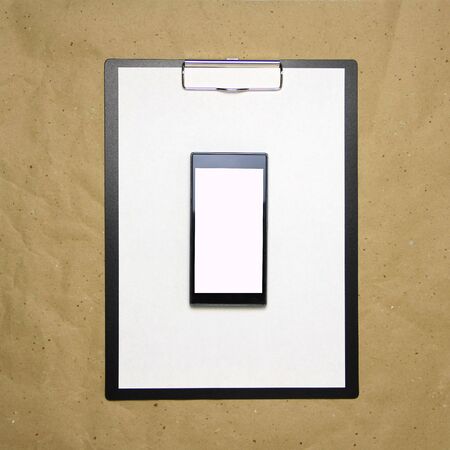 The tablet with a clip for the clip of paper with a white sheet a4 lies against the background of craft brown crumpled paper. Above is a mobile phone with a white screen. Place for text and layout for design.の写真素材