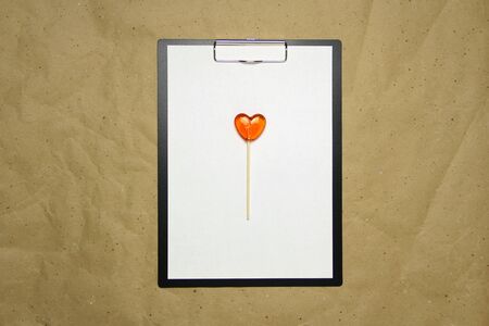 The tablet with a clip for the clip of paper with a white sheet a4 lies against the background of craft brown crumpled paper. On top lies a caramel lollipop of orange red in the shape of a heart. Place for text and layout for design.の写真素材