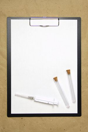 The tablet with a clip for the clip of paper with a white sheet a4 lies against the background of craft brown crumpled paper. On top is a medical syringe and two transparent empty tubes with stoppers. Place for text and layout for design.の写真素材
