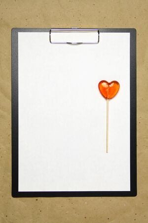 The tablet with a clip for the clip of paper with a white sheet a4 lies against the background of craft brown crumpled paper. On top lies a caramel lollipop of orange red in the shape of a heart. Place for text and layout for design.の写真素材