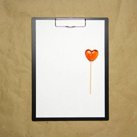 The tablet with a clip for the clip of paper with a white sheet a4 lies against the background of craft brown crumpled paper. On top lies a caramel lollipop of orange red in the shape of a heart. Place for text and layout for design.の写真素材