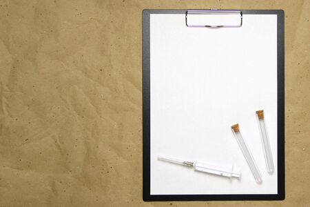 The tablet with a clip for the clip of paper with a white sheet a4 lies against the background of craft brown crumpled paper. On top is a medical syringe and two transparent empty tubes with stoppers. Place for text and layout for design.の写真素材