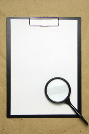 A tablet with a white sheet of A4 format with magnifier on a beige craft paper. Concept of analysis, study, attentive work. Stock photo with empty place for your text and design.の写真素材