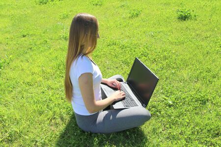 A beautiful young white girl in a white t-shirt and with long hair sitting on green grass, on the lawn and working behind a black laptop.の写真素材