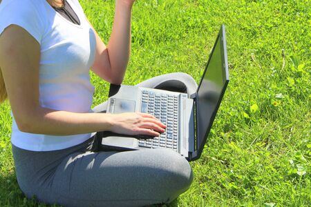 A beautiful young white girl in a white t-shirt and with long hair sitting on green grass, on the lawn and working behind a black laptop.の写真素材