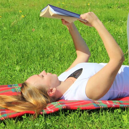 A beautiful young white girl in a white T-shirt and with long hair lies on a red plaid, on green grass, on the lawn and read book.の写真素材