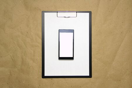 The tablet with a clip for the clip of paper with a white sheet a4 lies against the background of craft brown crumpled paper. Above is a mobile phone with a white screen. Place for text and layout for design.の写真素材