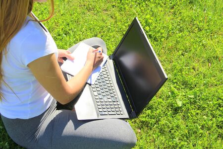 A beautiful young white girl in a white t-shirt and with long hair sitting on green grass, on the lawn and working behind a black laptop and writes with a pen in a notebook.の写真素材