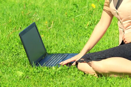 A beautiful young white girl in a pink jacket and black skirt and with long hair sitting on green grass, on the lawn and working behind a black laptop.の写真素材