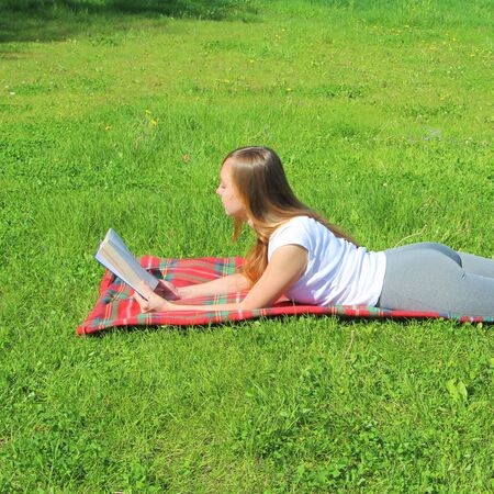 A beautiful young white girl in a white T-shirt and with long hair lies on a red plaid, on green grass, on the lawn and read book.の写真素材